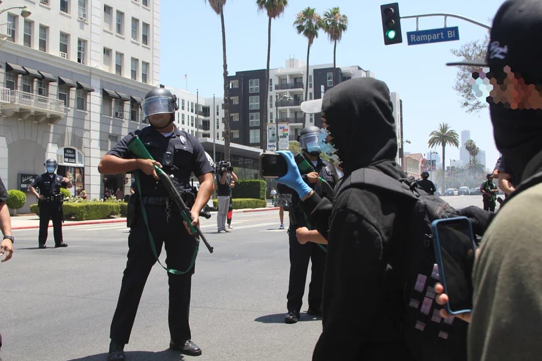 A masked activist wearing disposable gloves records LAPD officers in front of them on their smartphone. Officers can be seen holding green-marked bean-bag guns. The most visible officer in the image c