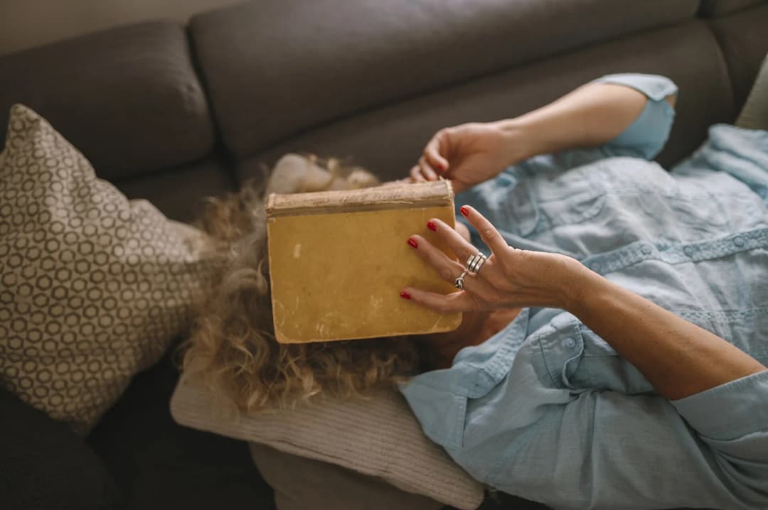 A person with curly hair is lying on a soft couch, holding a vintage book above their face. The atmosphere is relaxed, with decorative pillows and soft lighting.