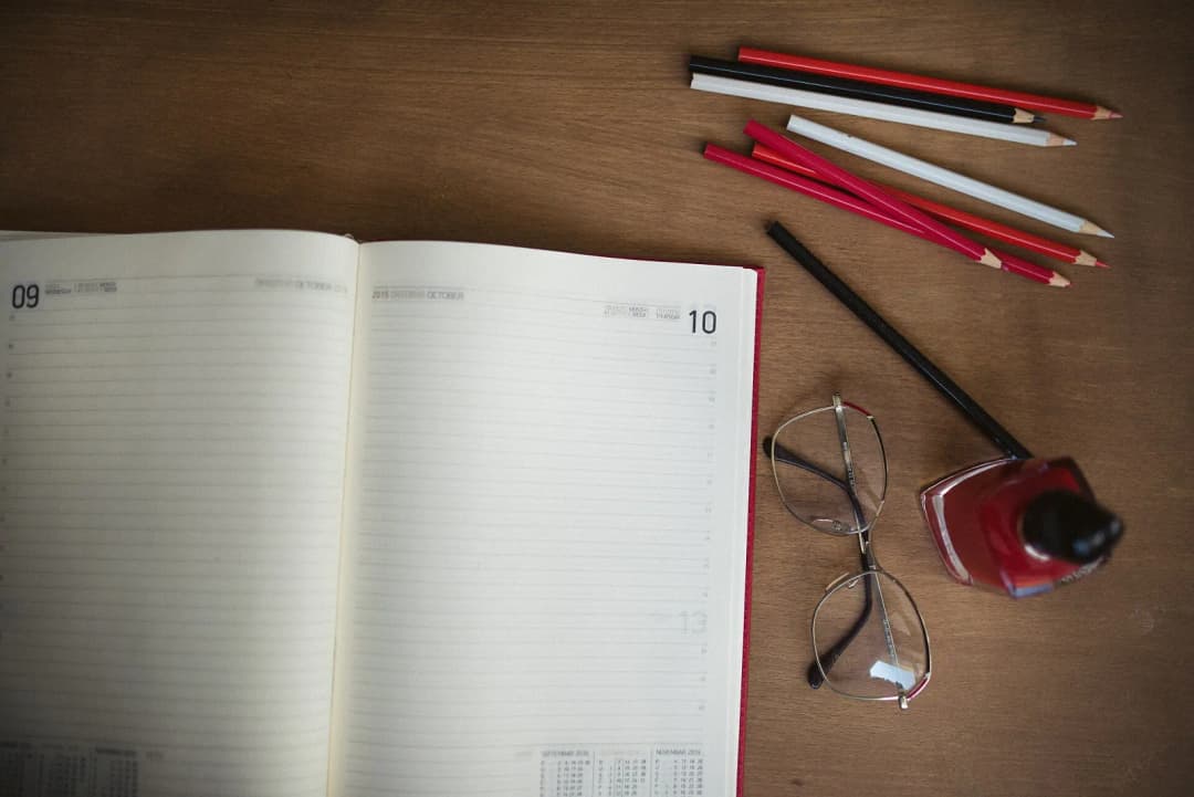 A planner is open to a new week with blank pages on a wooden desk. Nearby are colored pencils, glasses, and ink, suggesting an organized workspace for creative tasks.