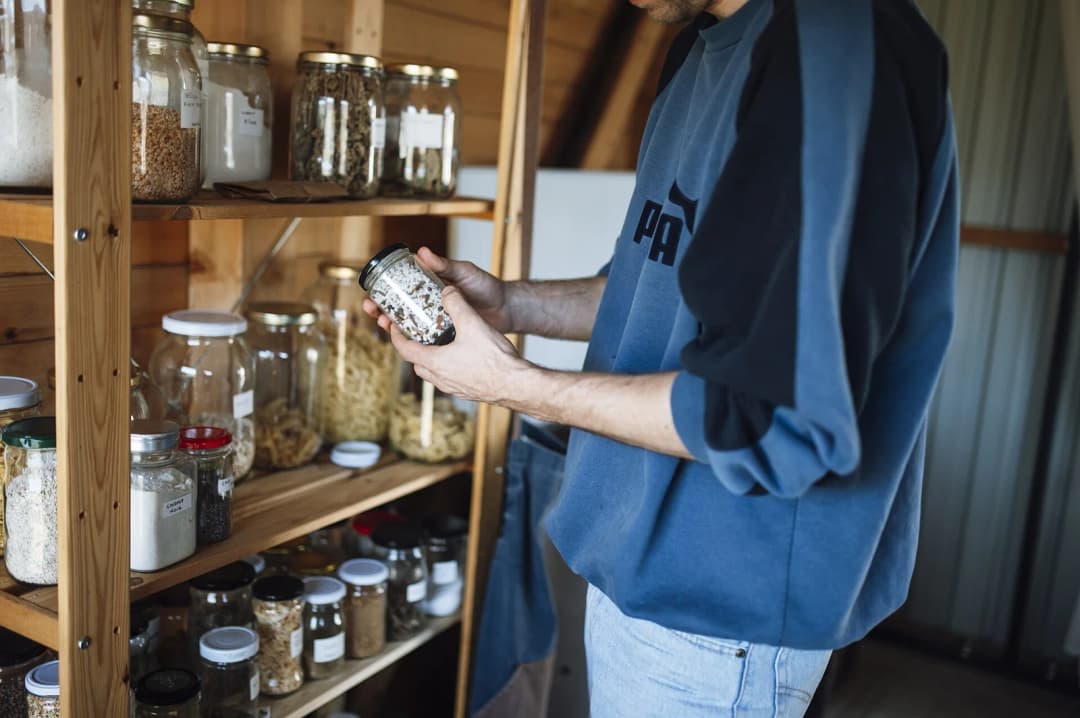 A person stands in a kitchen pantry looking closely at a jar filled with spices. The pantry has many jars arranged on wooden shelves. It is bright and organized.